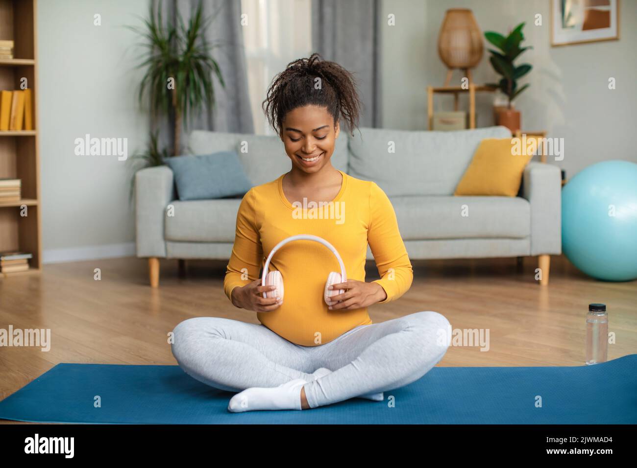 Garde d'enfants. Jeune femme afro-américaine joyeuse enceinte assise sur un tapis et appliquant des écouteurs sur le grand ventre Banque D'Images