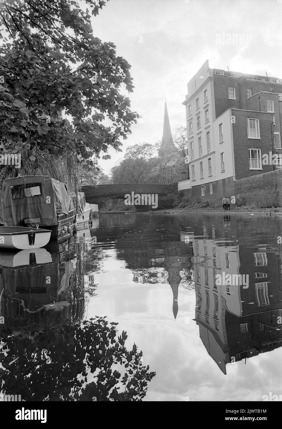 Londres, Angleterre, vers 1967. Vue depuis le canal Regent's près de Camden, Londres. Un bâtiment se trouve devant vous, sur la place St Marc et le pont Regent's Park Road. Au loin se trouve la flèche de l’église Saint-Marc. Des bateaux sont amarrés sur le canal et un couple marche le long du chemin de halage. Banque D'Images
