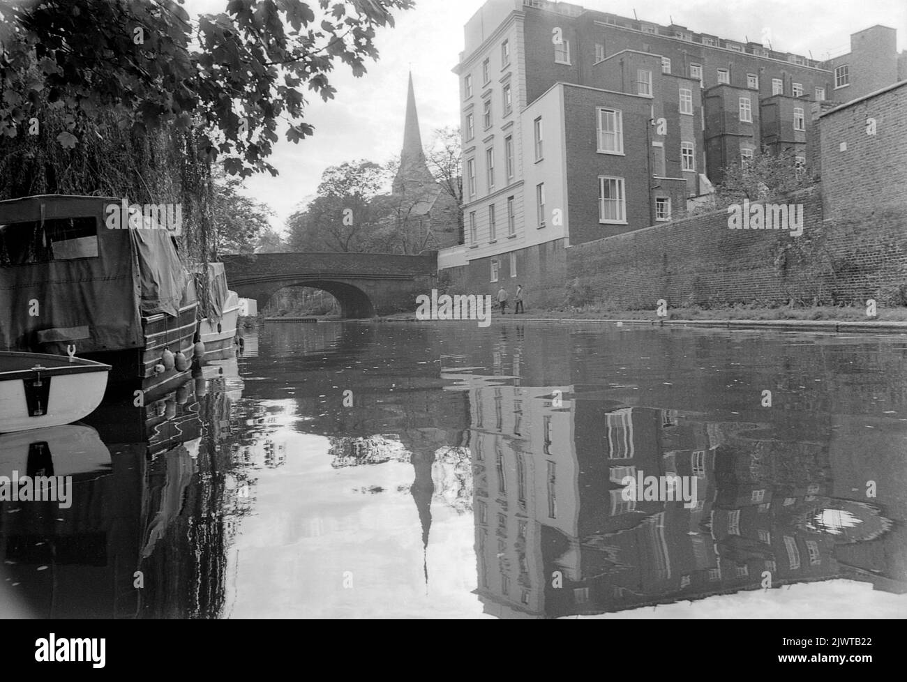 Londres, Angleterre, vers 1967. Vue depuis le canal Regent's près de Camden, Londres. Un bâtiment se trouve devant vous, sur la place St Marc et le pont Regent's Park Road. Au loin se trouve la flèche de l’église Saint-Marc. Des bateaux sont amarrés sur le canal et un couple marche le long du chemin de halage. Banque D'Images