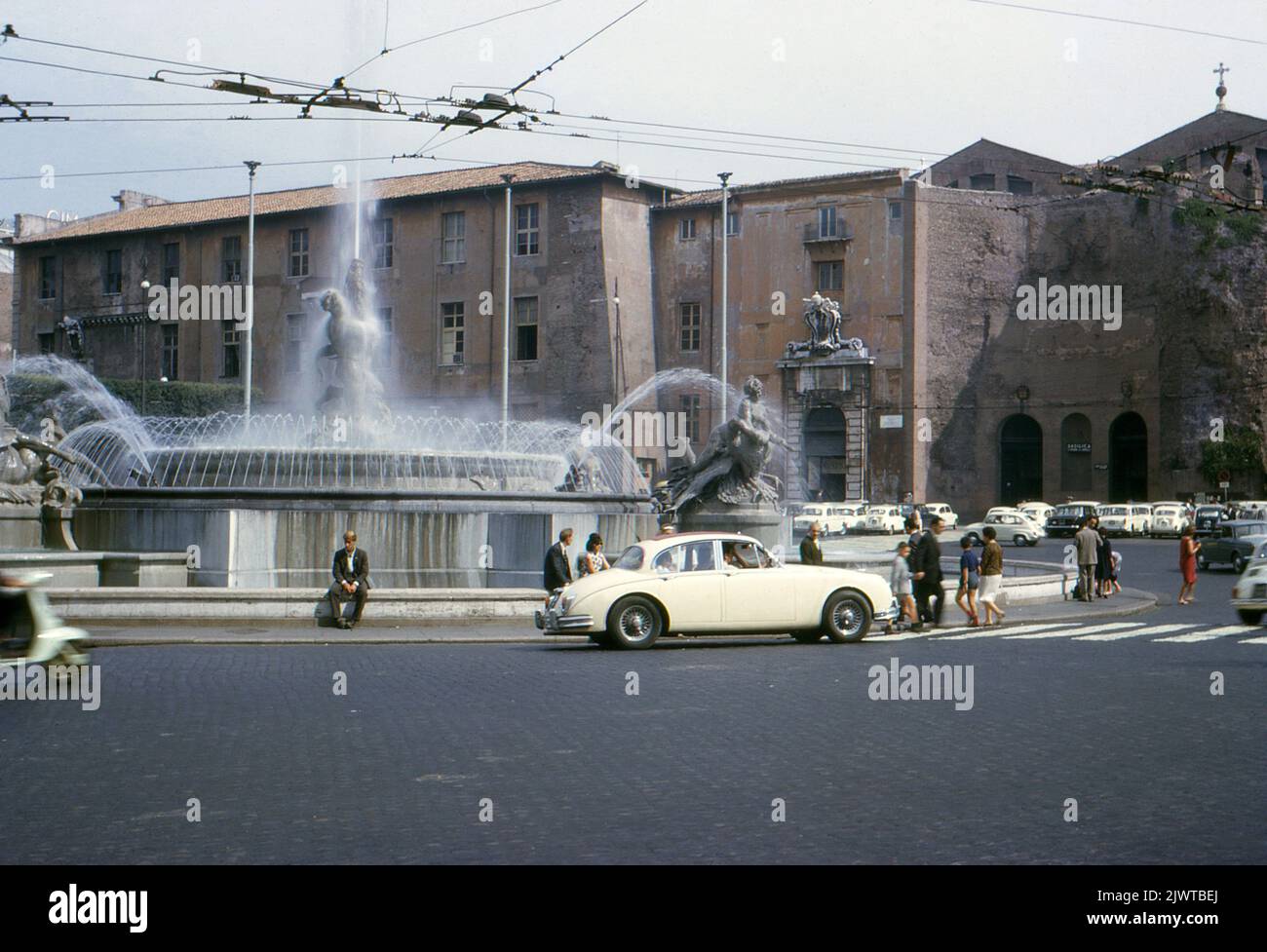 Rome, 1963. Vue sur la fontaine des Naiads, située au centre de la Piazza della Repubblica, sur la colline Viminal. La fontaine a été créée par l'architecte Alessandro Guerrieri en 1888. Des sculptures en bronze de naiads et d'un autre de Dieu glaucus ont été créées par Mario Rutelli et ajoutées en 1901 et 1912. Une voiture Jaguar Mk.II attend tandis que des piétons traversent la route en face. Derrière la fontaine se trouve l'entrée de la basilique Sainte-Marie des Anges et des Martyrs. Banque D'Images