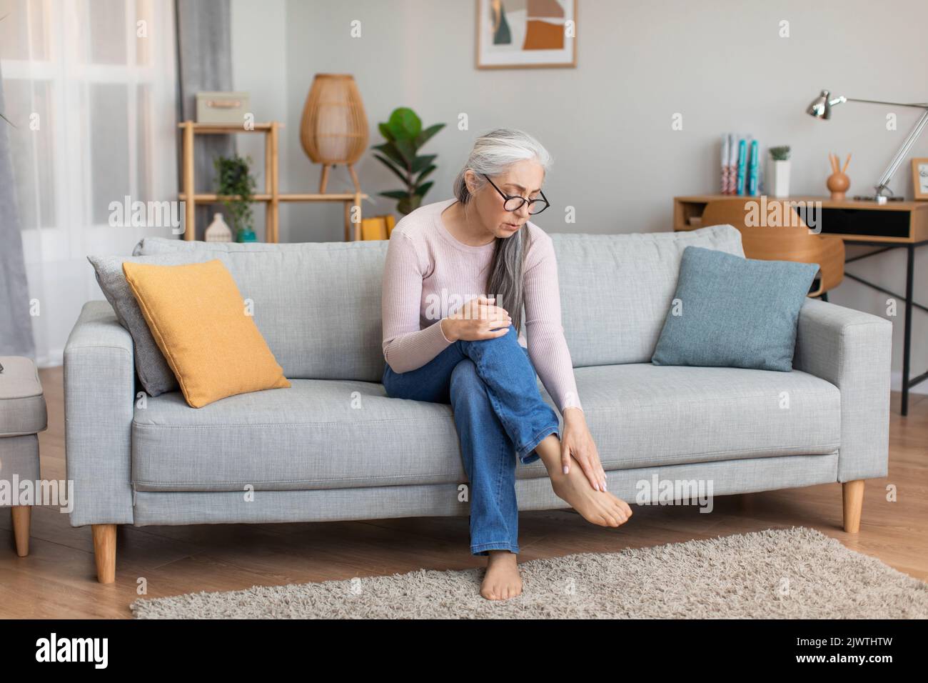 Triste caucasien âgé gris-cheveux femme dans les verres assis sur le canapé touche la jambe, souffrant de paine Banque D'Images