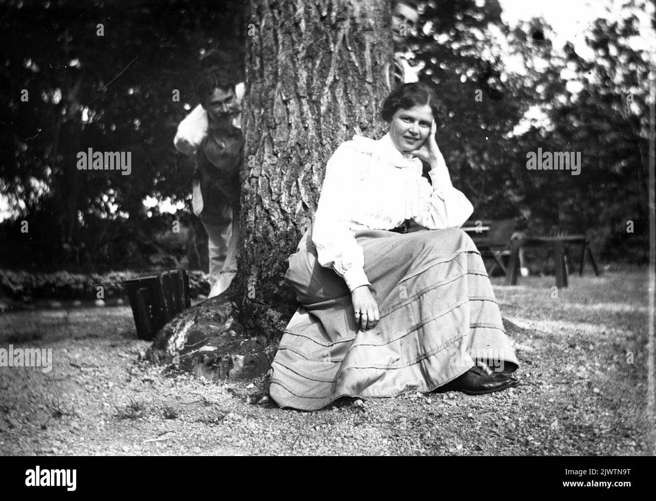 Femme assise sous l'arbre, les hommes regardent vers l'avant derrière l'arbre Kvinna sitter sous trädet, männen tittar fram bakom trädet Banque D'Images