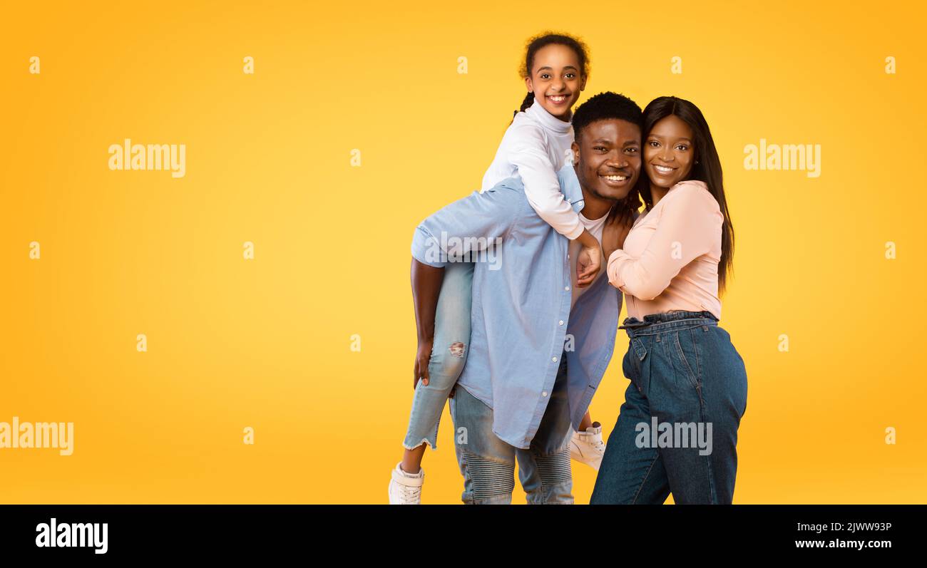 Jeune famille afro-américaine heureuse posant, homme donnant le dos de piggy pour sa fille, fond jaune, espace libre Banque D'Images