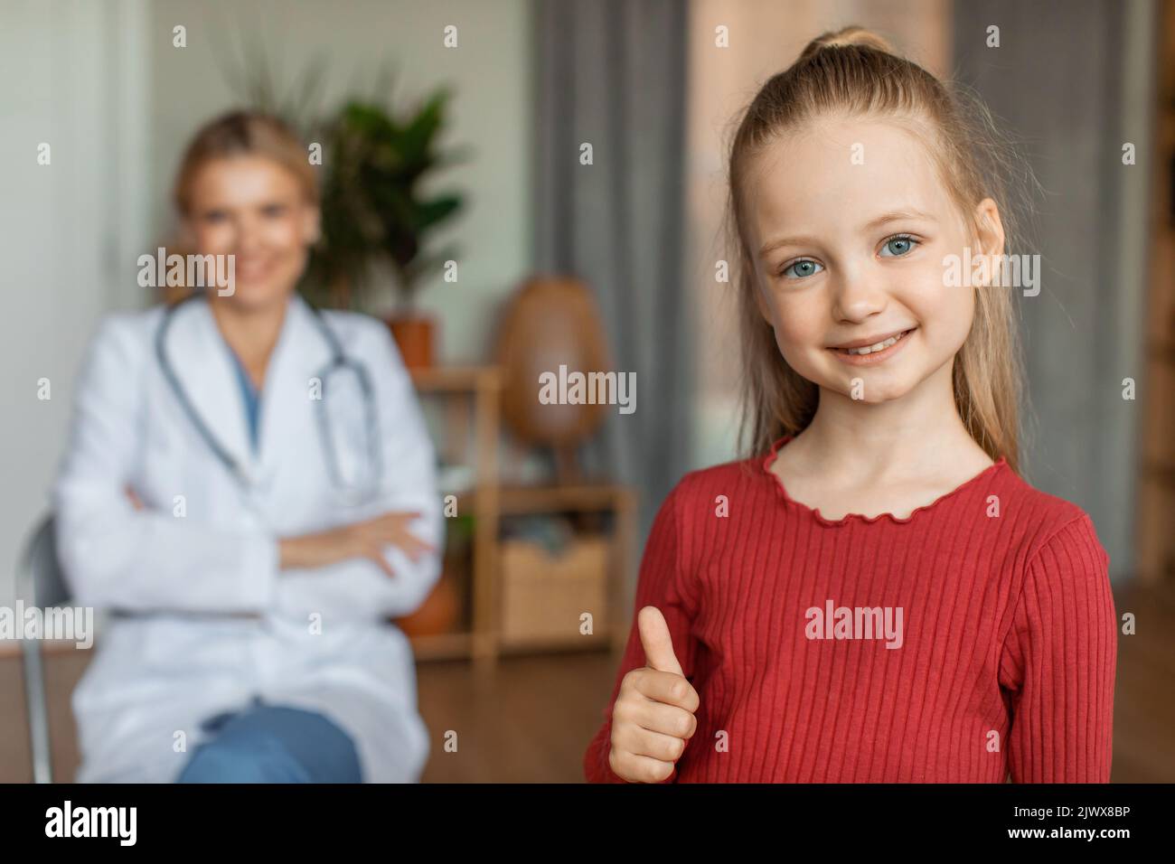 Bonne petite fille à la clinique pédiatre avec une femme médecin sur fond souriant à l'appareil photo et montrant le pouce vers le haut Banque D'Images