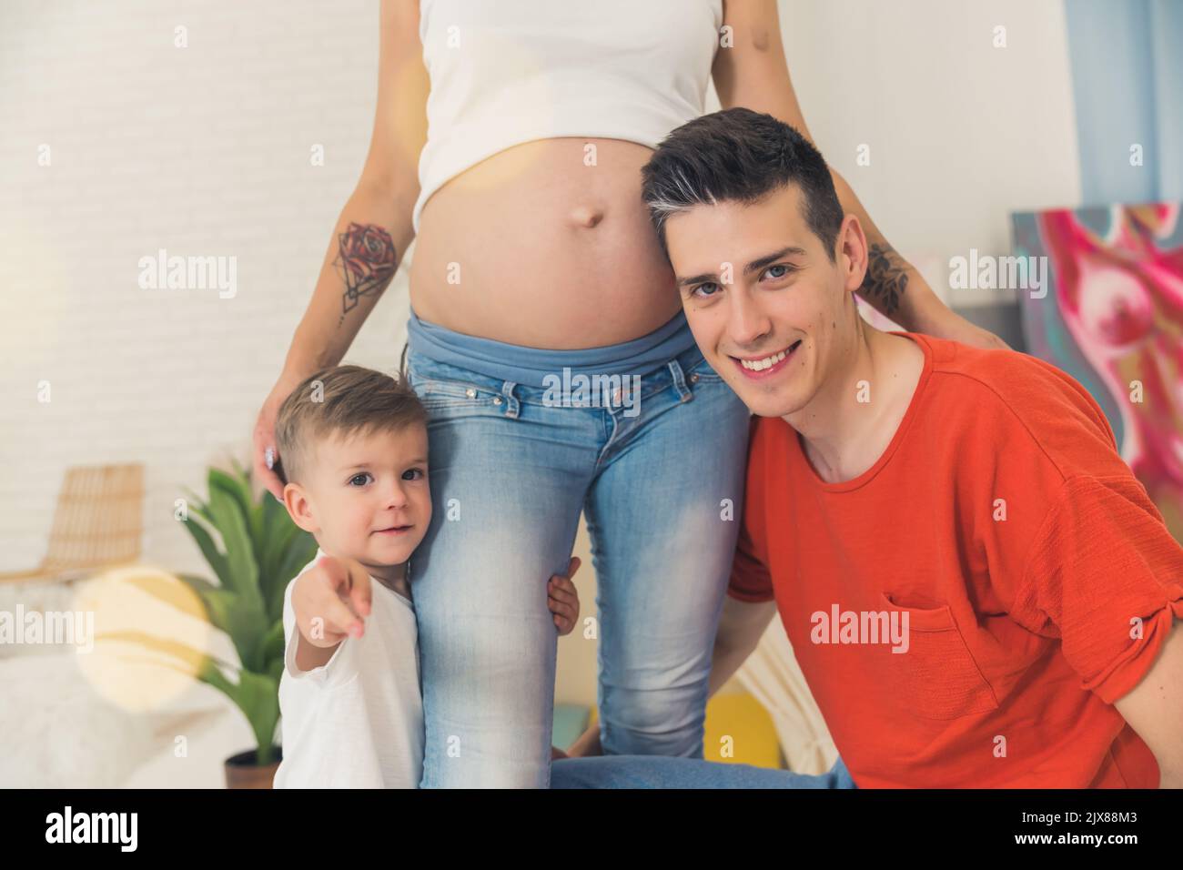 La jolie famille européenne s'agrandit. Homme caucasien à poil sombre et son jeune garçon adorable tenant les jambes de la femme caucasienne enceinte en mamans jeans. Photo de haute qualité Banque D'Images