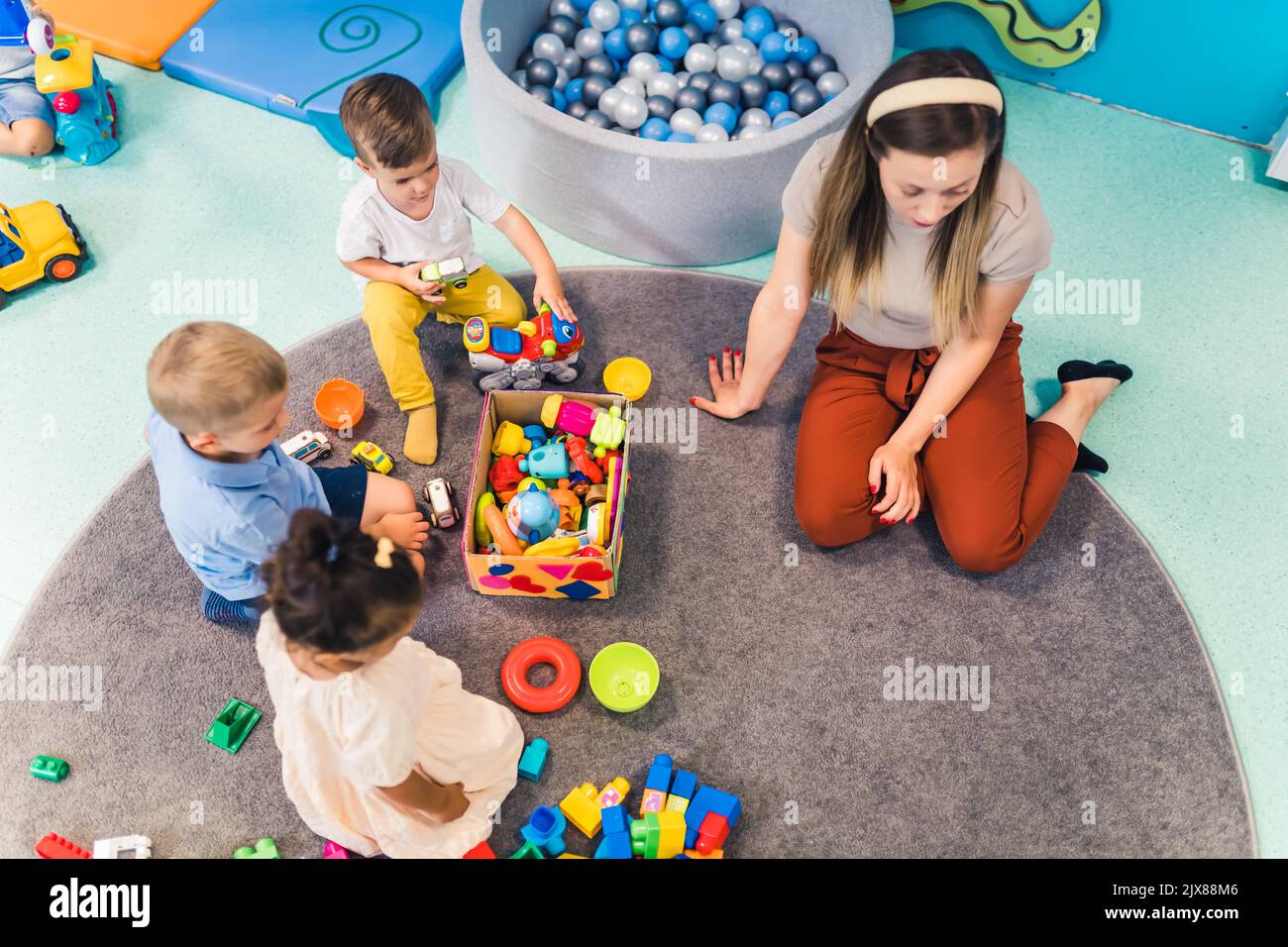 Jeux à l'école maternelle. Les tout-petits avec leur professeur assis sur le sol et jouant avec des blocs de construction, des voitures colorées et d'autres jouets. Photo de haute qualité Banque D'Images