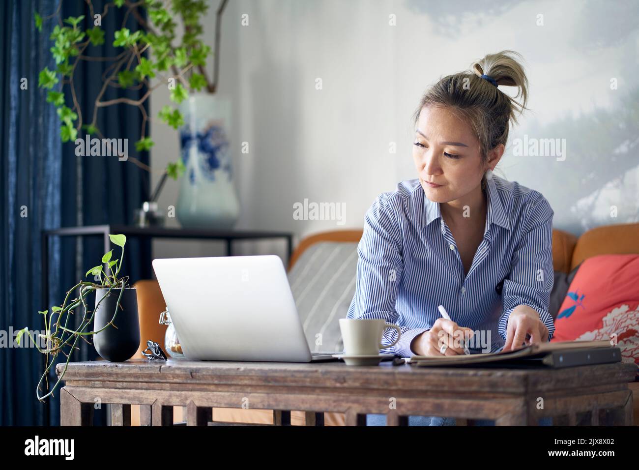 femme asiatique professionnelle mûre travaillant à la maison en prenant des notes tout en regardant un ordinateur portable Banque D'Images