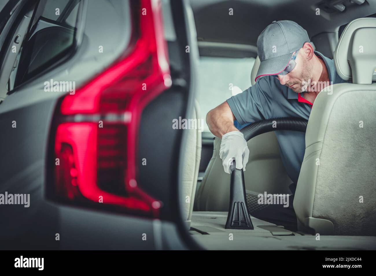 Le détaleur automatique caucasien aspire avec précision les sièges arrière rabattus pendant le service de nettoyage intérieur de véhicule de qualité supérieure. Banque D'Images