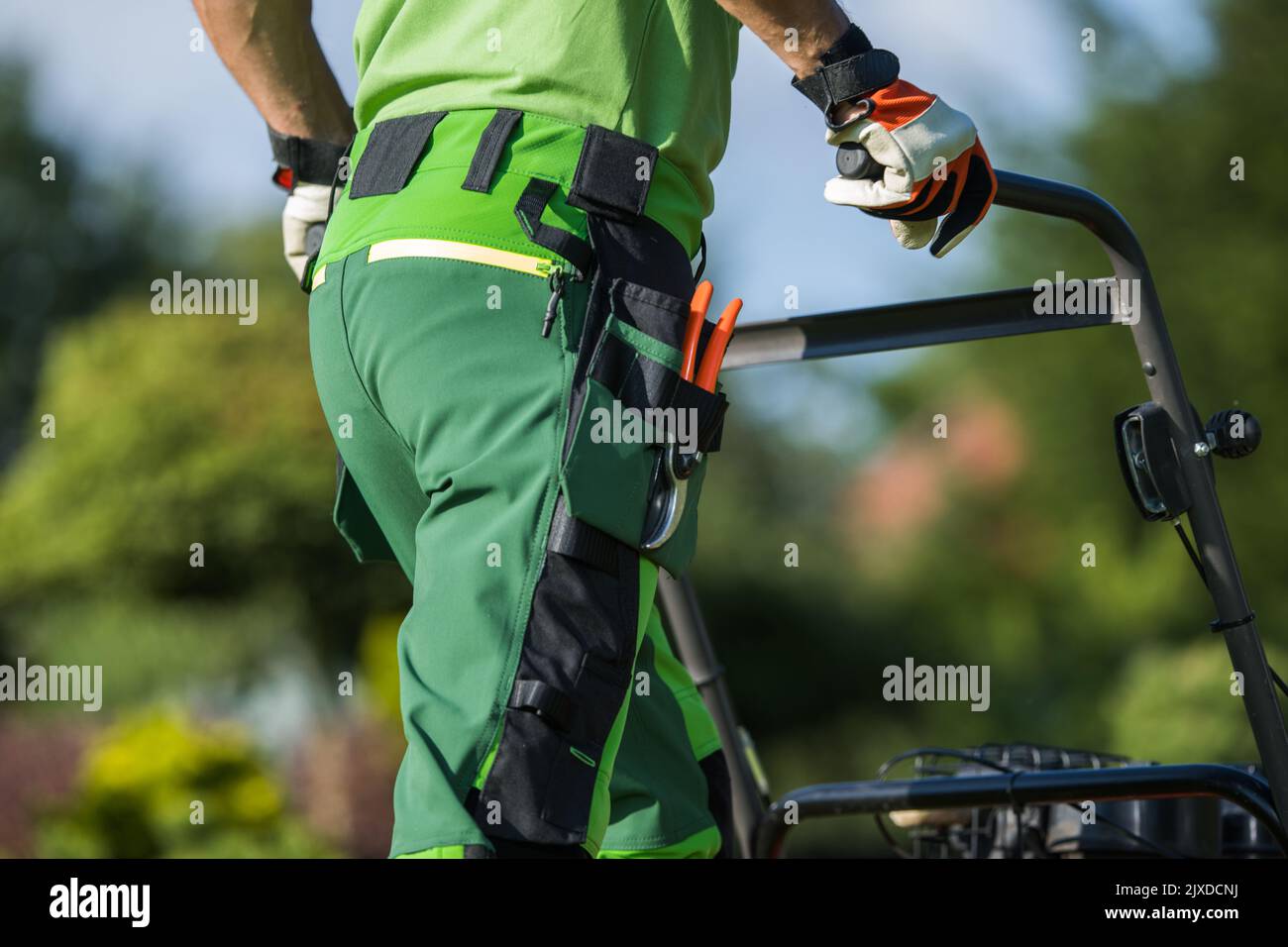 Jardinier caucasien debout à l'envers en uniforme de travail avec des ciseaux de jardin dans la poche poussant la tondeuse de pelouse. Equipement d'aménagement paysager professionnel Banque D'Images