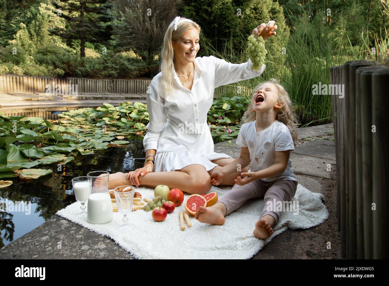 Drôle de famille assis sur un plat avec des fruits, un pot de lait sur le bord de l'étang. Jeune femme tenant des raisins sur une petite fille. Banque D'Images