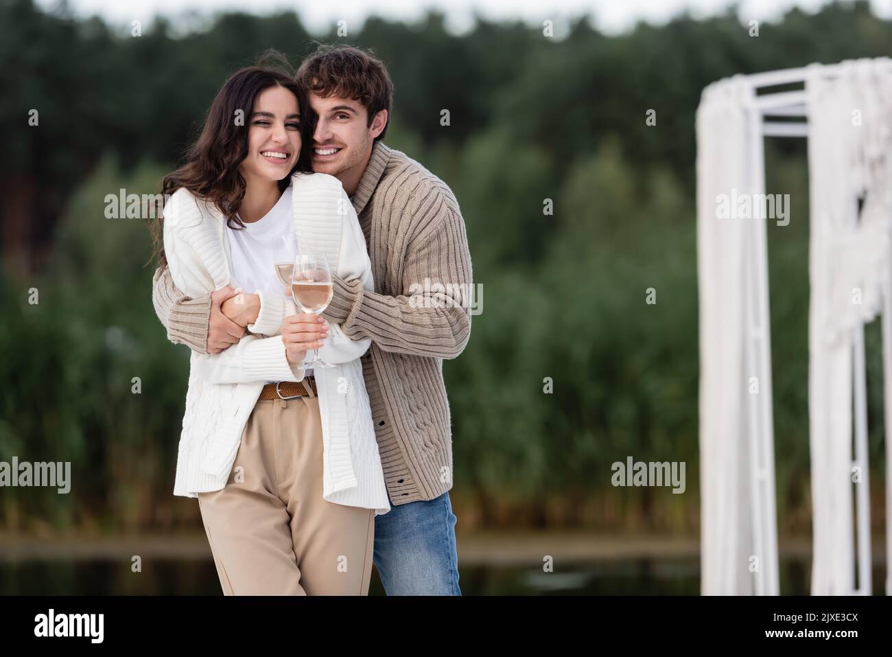 Couple souriant dans cardigans embrassant et tenant le vin à l'extérieur, image de stock Banque D'Images