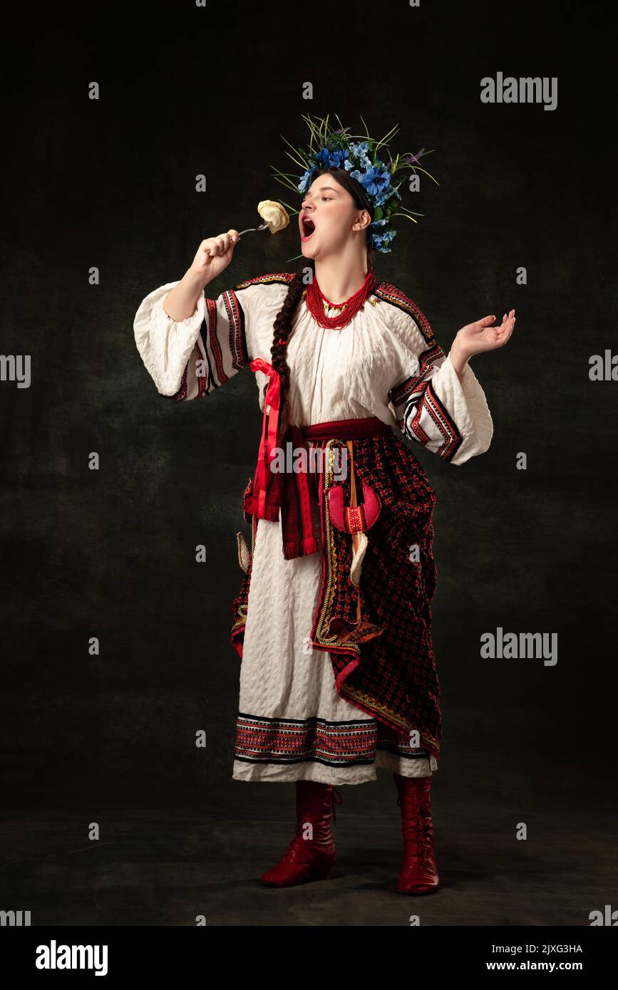 Portrait créatif de la belle femme ukrainienne portant un costume traditionnel folklorique dégustation varenik isolé sur fond sombre. Traditions, nationales Banque D'Images