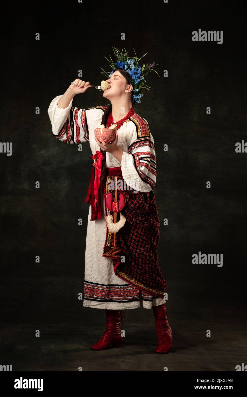 Profitez-en. Portrait créatif de la belle femme ukrainienne portant un costume traditionnel folklorique dégustation varenik isolé sur fond sombre. Banque D'Images