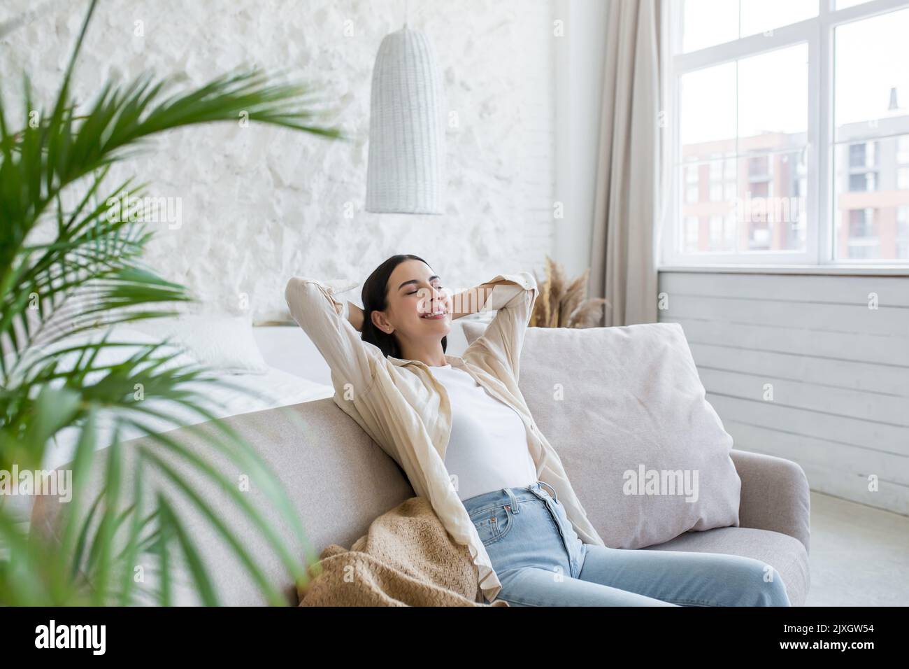 Une jeune belle femme d'affaires se repose sur le canapé après une journée de travail réussie. Pause. Elle s'assoit contentement à la maison près de la fenêtre avec ses yeux fermés, les mains derrière sa tête, souriant. Banque D'Images