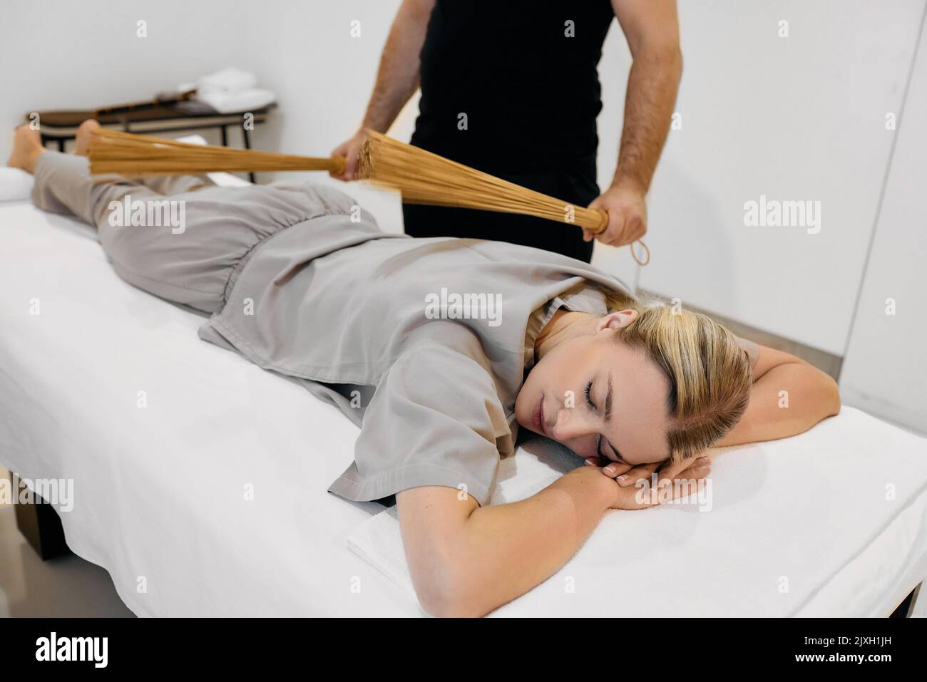 Jolie femme bénéficiant d'un massage avec des balais en bambou couchés sur une table de massage blanche avec un masseur. Massage thérapeutique avec bâtonnets de bambou Banque D'Images