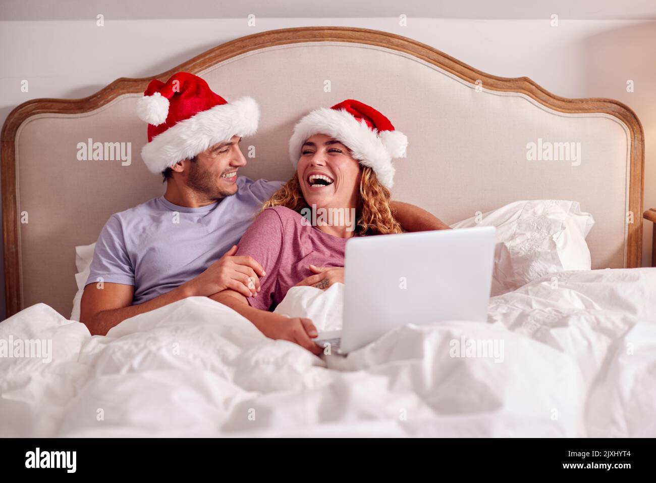 Couple à Noël allongé dans le lit à la maison faire appel vidéo sur ordinateur portable portant Santa Hats Banque D'Images