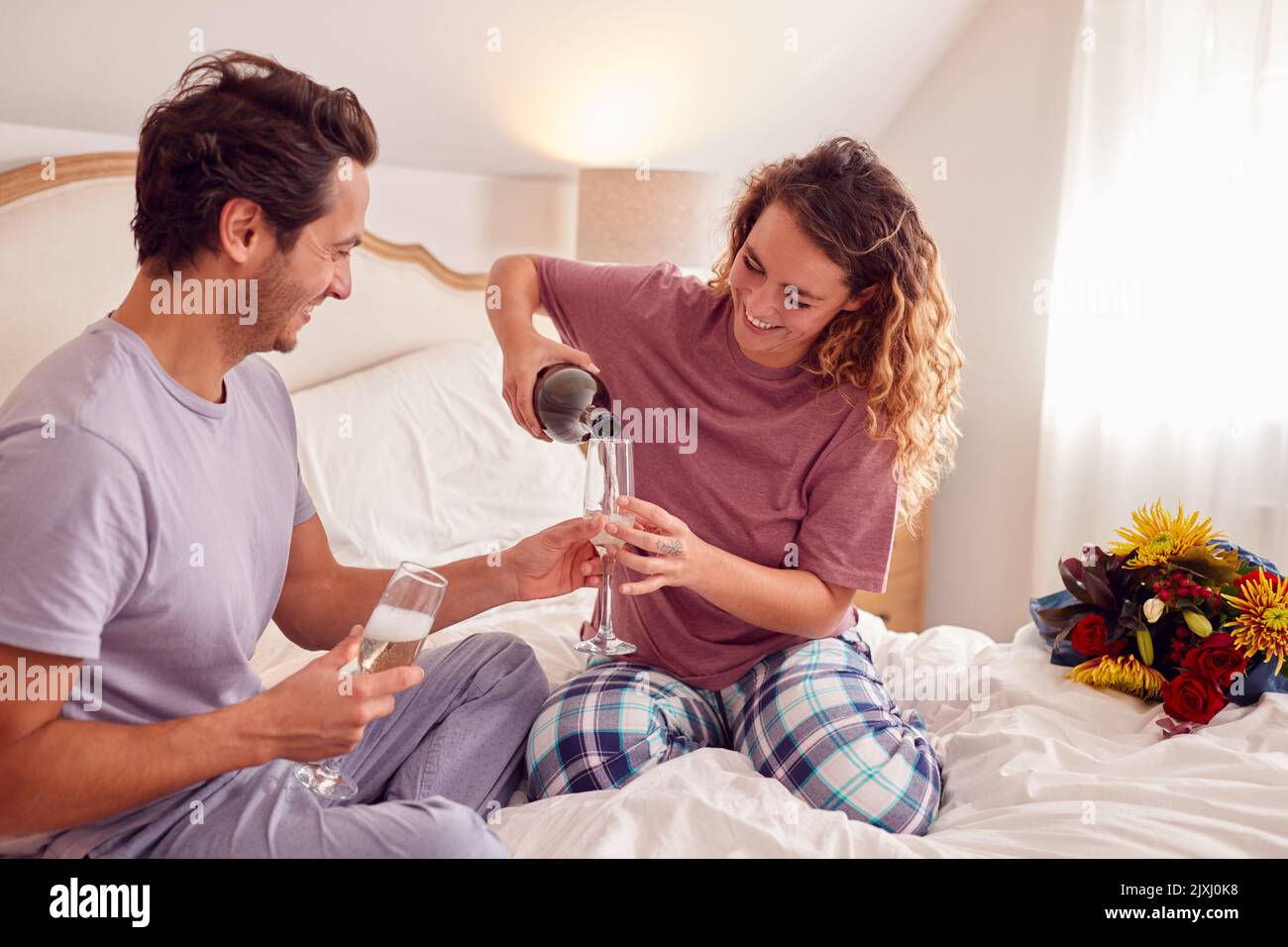 Couple aimant portant un pyjama dans le lit à la maison célébrant son anniversaire ou son anniversaire avec champagne Banque D'Images
