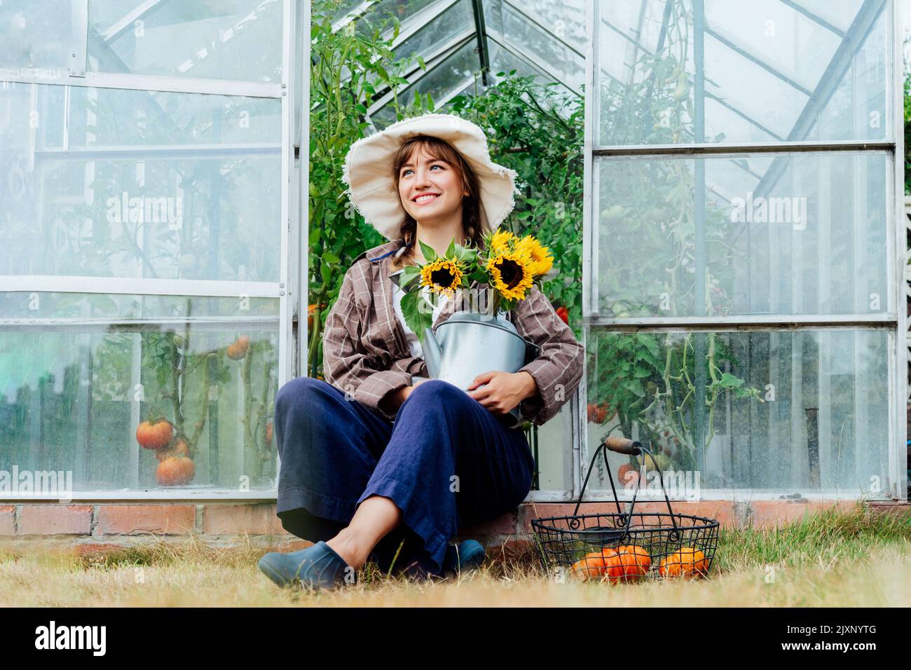 Une jeune femme paysanne souriante tient un arrosoir avec un bouquet de tournesols frais, assis près d'une serre avec des tomates fraîchement cueillies dans un panier. Cottagecore Banque D'Images Une jeune femme paysanne souriante tient un arrosoir avec un bouquet de tournesols frais, assis près d'une serre avec des tomates fraîchement cueillies dans un panier. Cottagecore Banque D'Images