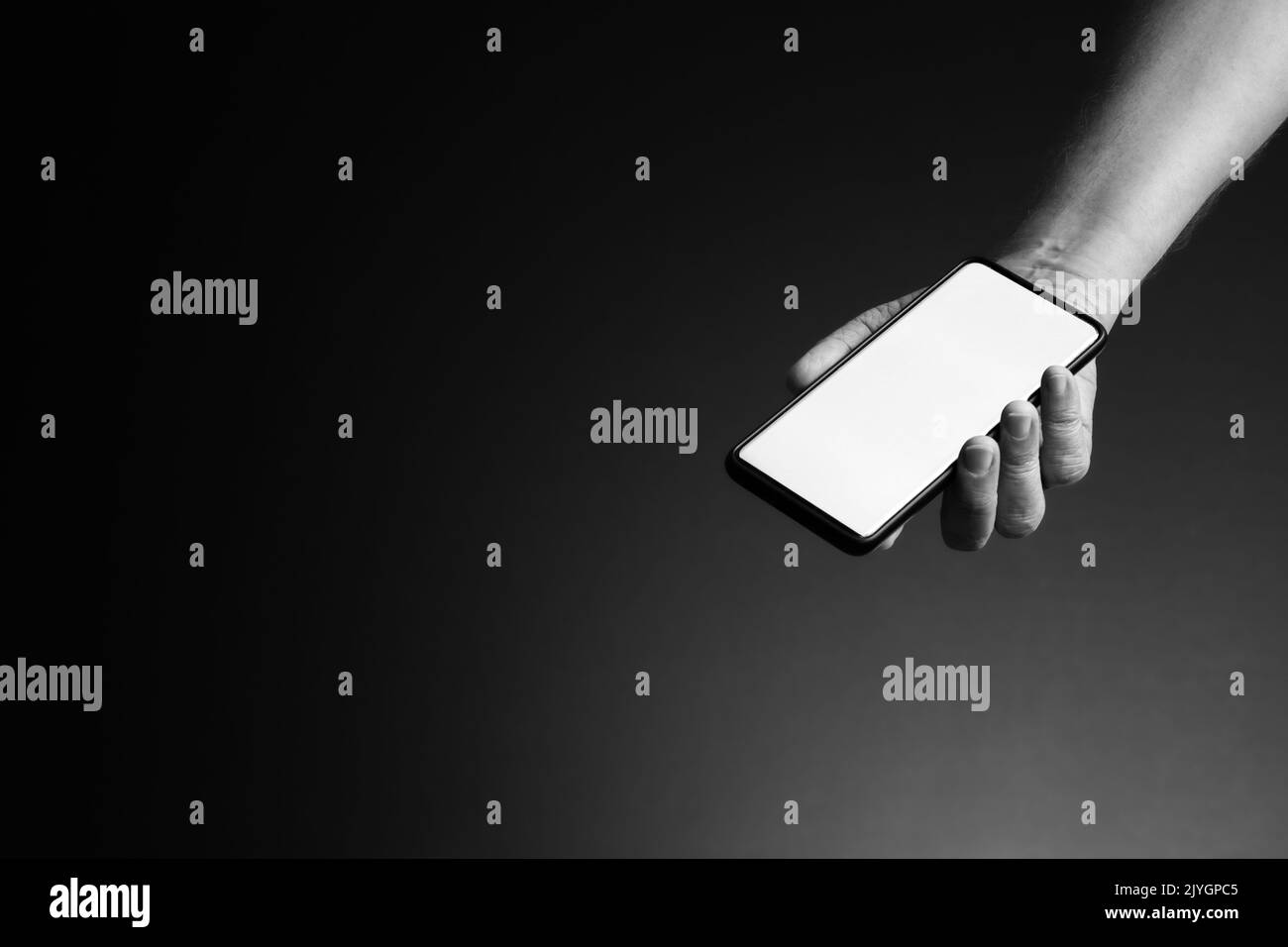 Image en noir et blanc de la main de l'homme étirée tenant le smartphone dans la paume avec écran blanc vierge isolé sur fond sombre avec espace de copie Banque D'Images