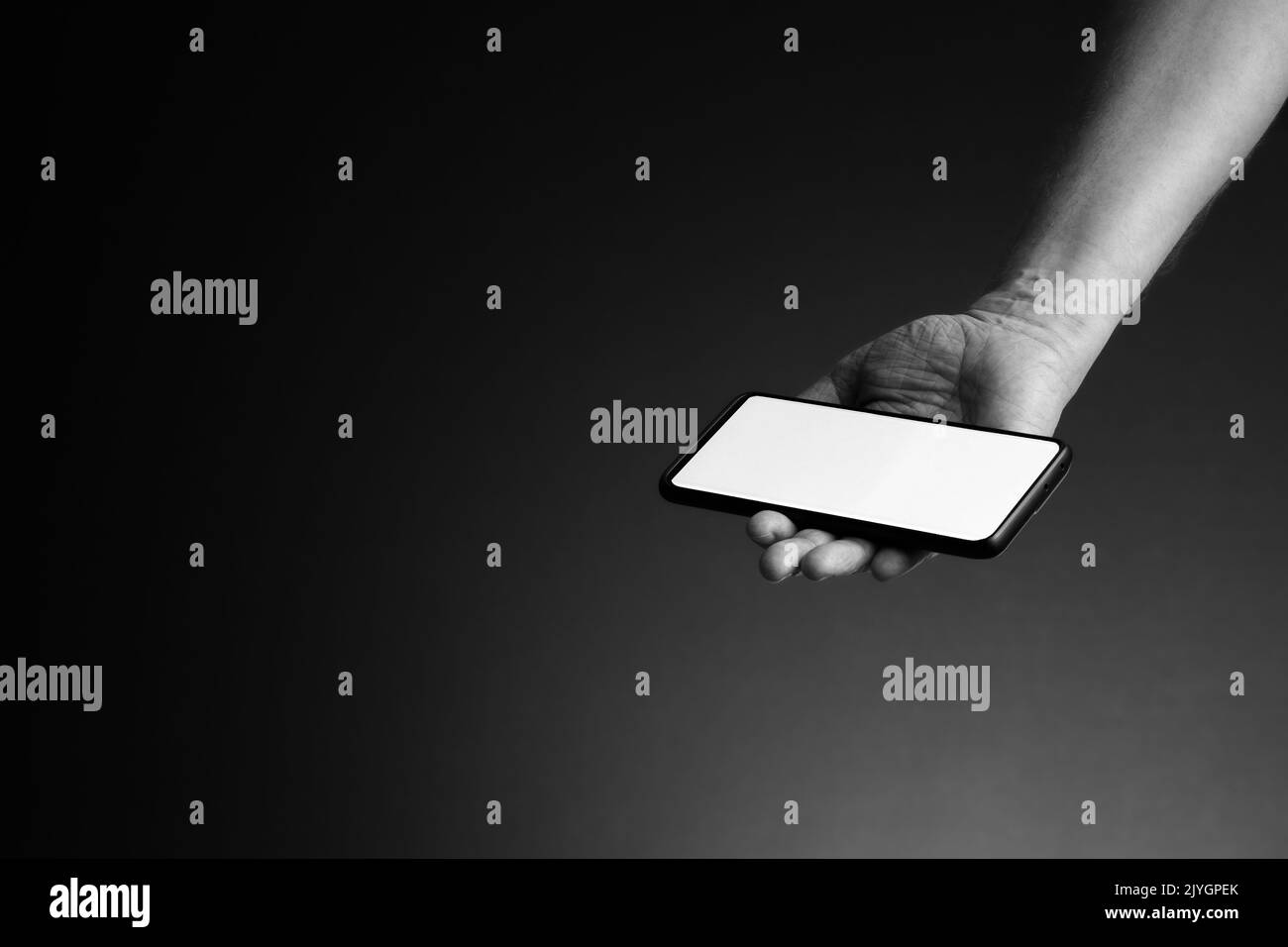 Image en noir et blanc de la main de l'homme étirée tenant le smartphone dans la paume avec écran blanc vierge isolé sur fond sombre avec espace de copie Banque D'Images