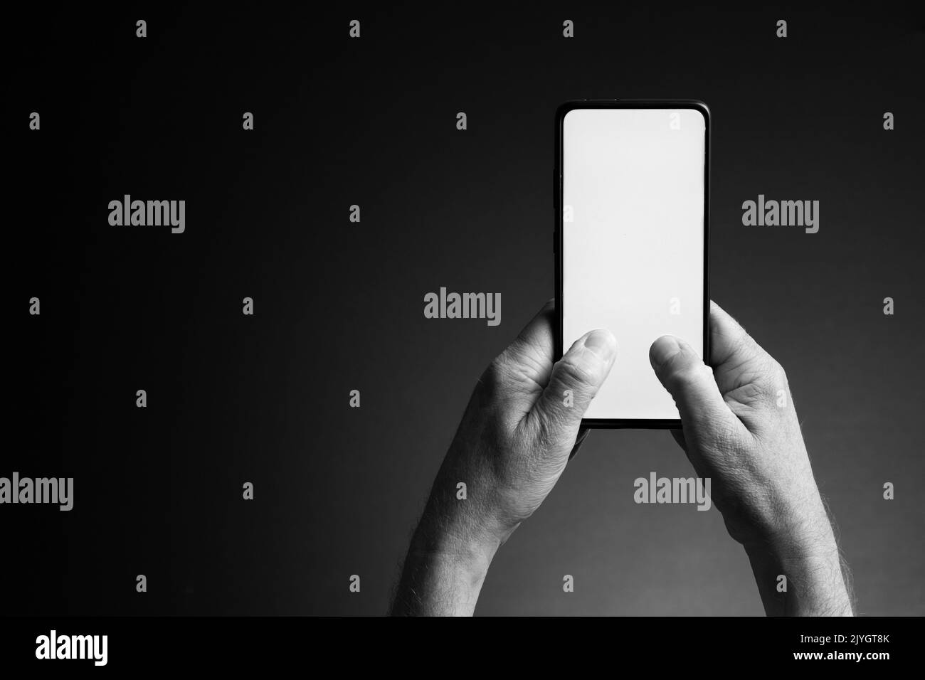 Image en noir et blanc des mains de l'homme tenant le smartphone et l'envoi de SMS avec écran blanc vierge isolé sur fond sombre avec espace de copie Banque D'Images