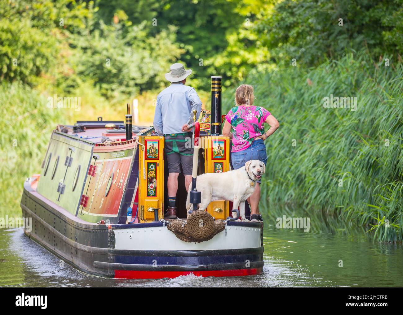 Vue arrière d'un couple et de son chien à bord d'un bateau sur le canal le jour de l'été. Le chien du labrador regarde en arrière sur le canal. Banque D'Images