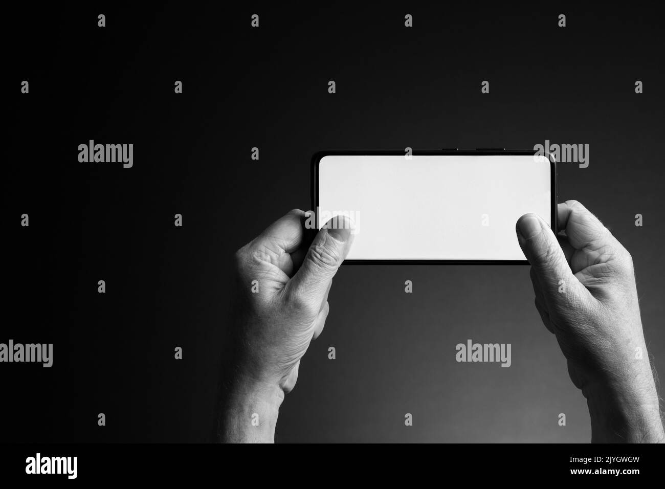 Image en noir et blanc des mains de l'homme tenant le smartphone horizontalement pour envoyer des SMS ou jouer avec un écran blanc vierge isolé sur fond sombre avec copie Banque D'Images