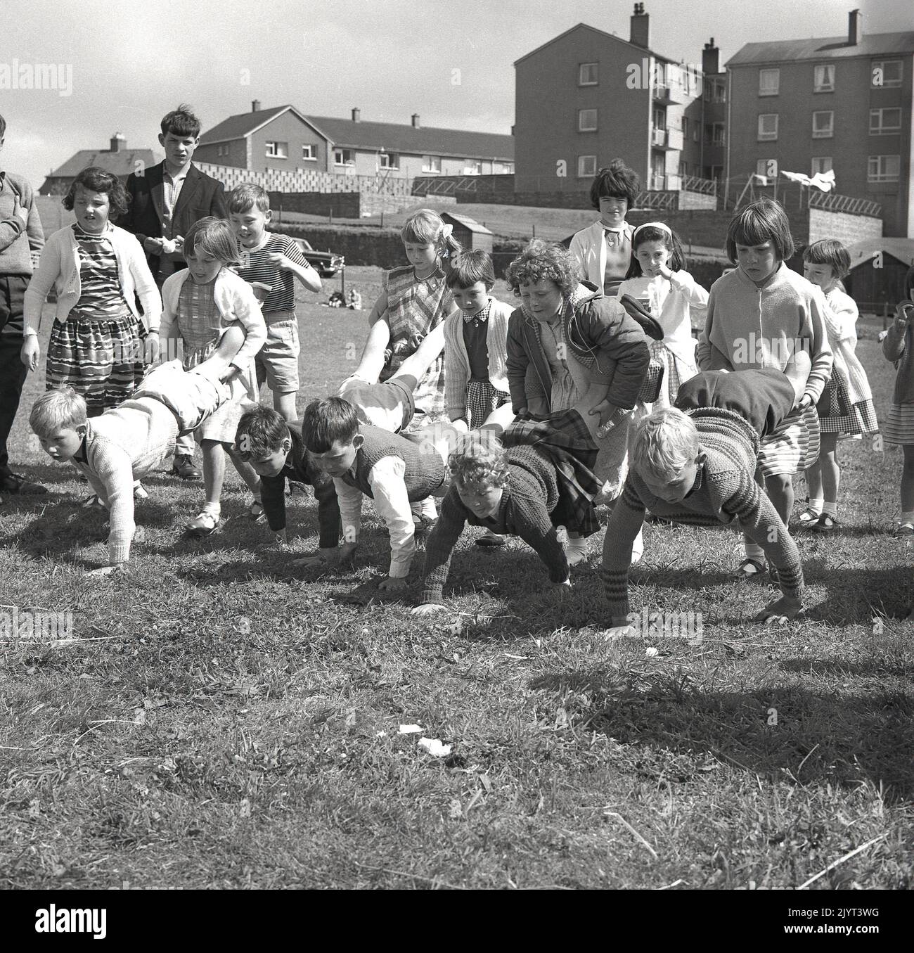 1965, historique, n. queensferry gala jour, les enfants prêts à commencer un match, une course roue-barrow dehors sur l'herbe dans le domaine d'un logement à North Queensferry, Fife, Écosse, Royaume-Uni, les jeunes filles tenant les jambes des garçons! Banque D'Images