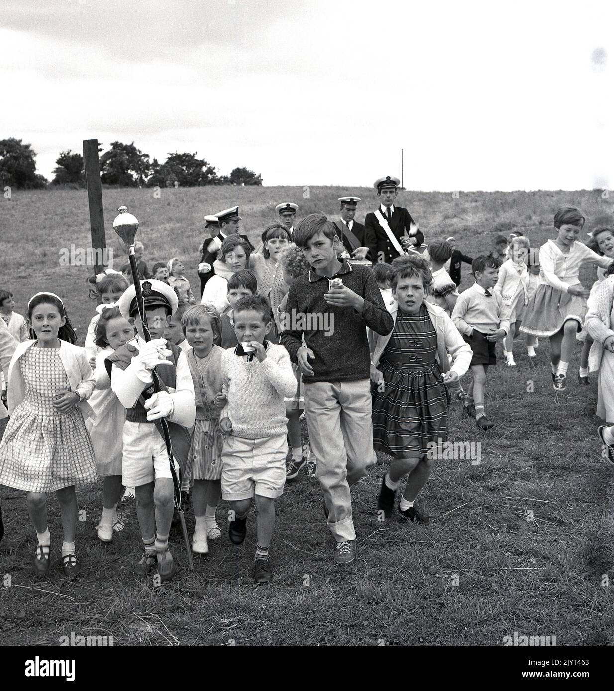 1965, historique, enfants à l'extérieur dans un champ près d'un domaine de logement prenant part au gala du North Queensferry, Fife, Écosse, Royaume-Uni, les enfants suivent un garçon habillé comme un tambour majeur tenant un mace. Banque D'Images