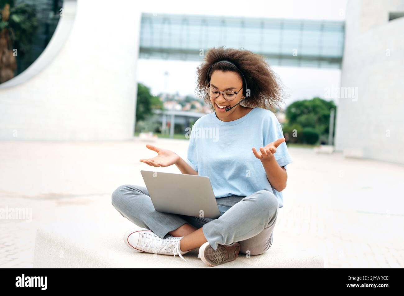 Une jeune femme afro-américaine satisfaite, aux cheveux mauriquement, avec micro-casque, travailleur de l'entreprise, consultation en ligne, négociations avec des collègues, assis à l'extérieur, utilisation d'un ordinateur portable, conversation vidéo, sourire Banque D'Images