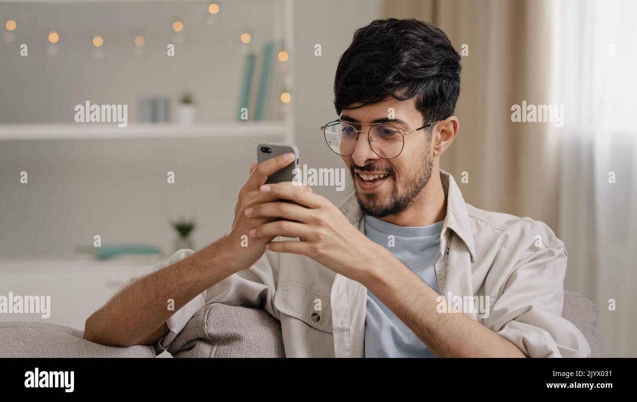 Jeune 30s arabe barbu homme millénaire millenny en lunettes assis à la maison sur un canapé regardant le téléphone mobile faire oui geste obtenir l'offre gagnant bonne nouvelle Banque D'Images