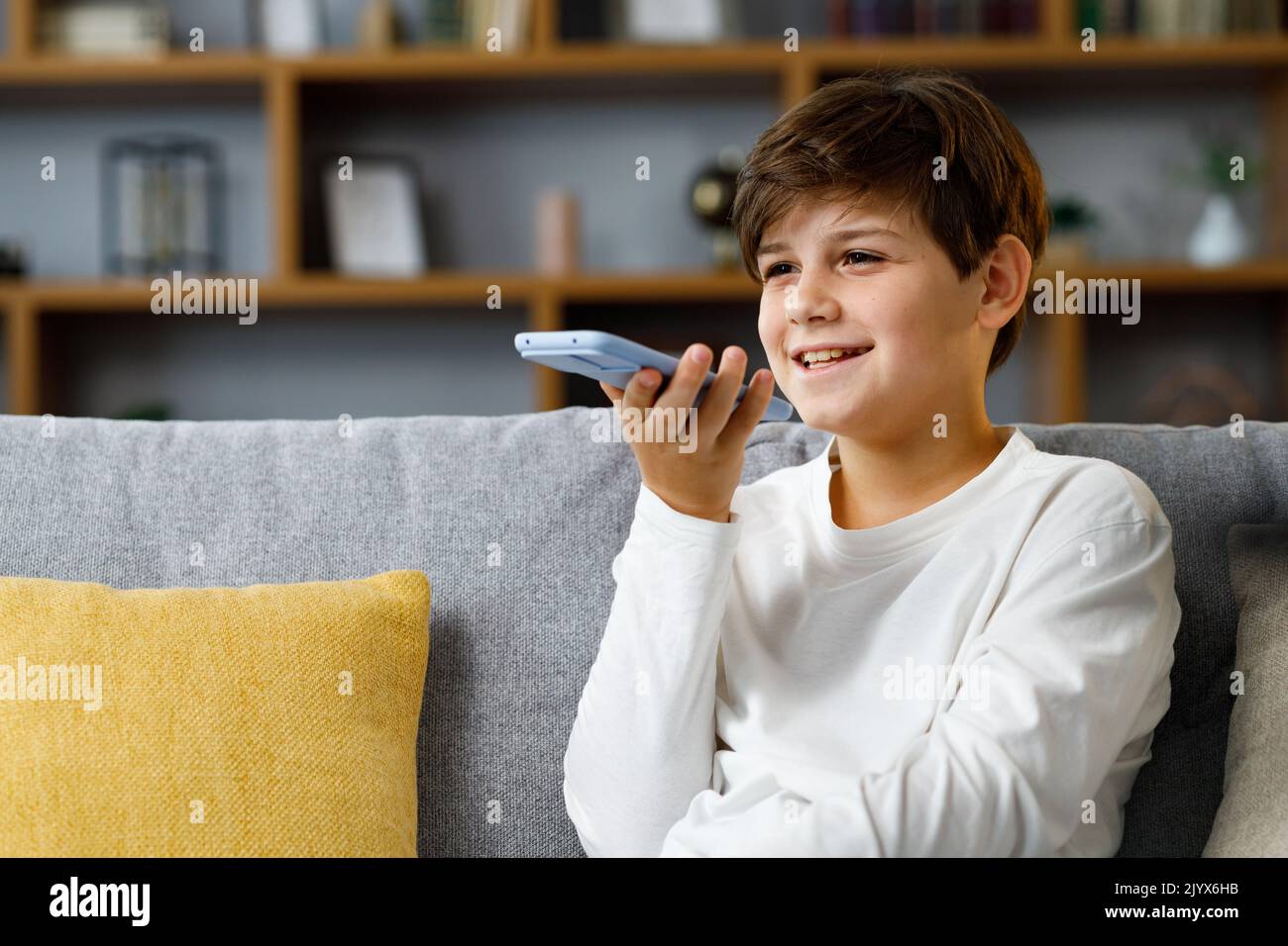 Jeune garçon mignon enregistrant un message vocal sur un smartphone. Adolescent souriant à l'aide de l'application d'assistant virtuel assis à l'entraîneur à la maison. Reconnaissance vocale, audio Banque D'Images