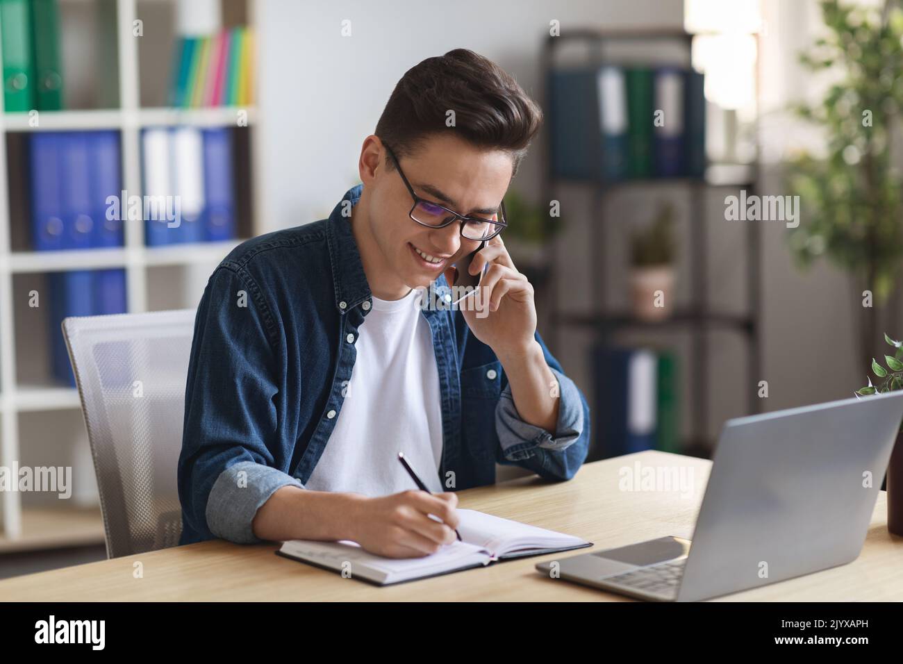Jeune responsable de bureau parlant sur un téléphone mobile et prenant des notes sur le Bloc-notes Banque D'Images
