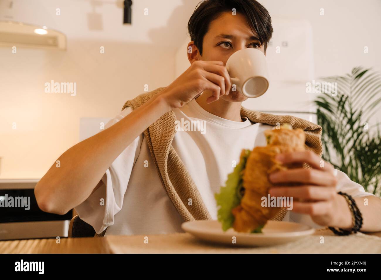 Un garçon de brunette asiatique mangeant un sandwich et buvant du café dans la cuisine à la maison Banque D'Images