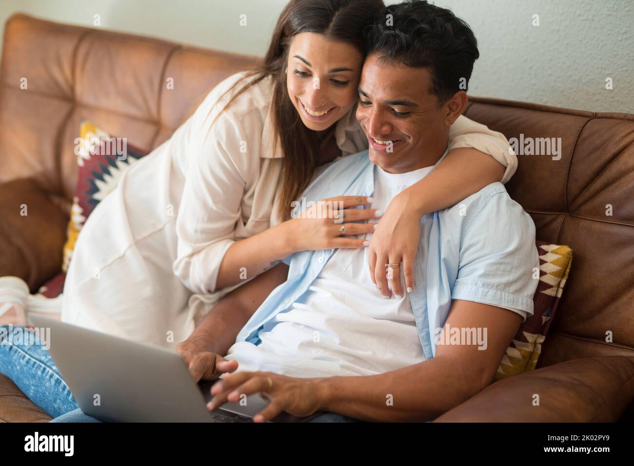 Joyeux couple interracial de race blanche et noire avec une jeune femme qui embrasse son petit ami pendant qu'il travaille sur un ordinateur portable. Relation et amitié dans le travail intelligent activité à l'intérieur à la maison assis sur le canapé Banque D'Images