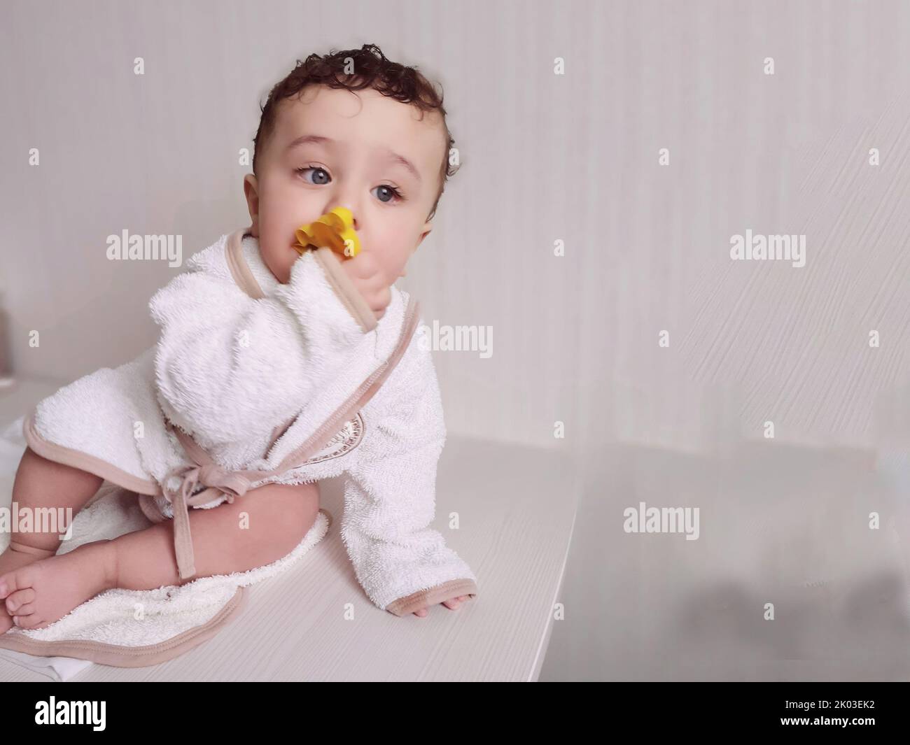 Un petit garçon avec des cheveux blonds et des yeux dans un peignoir est assis sur une table. Banque D'Images