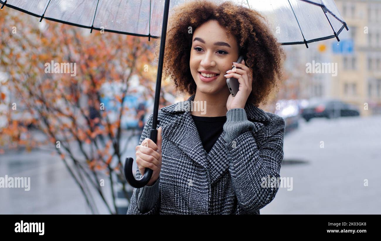 Bonne active amicale mauriquement cheveux fille afro-américaine avec parapluie transparent dans la ville dans le vent pluvieux temps répondre à l'appel parlant Banque D'Images