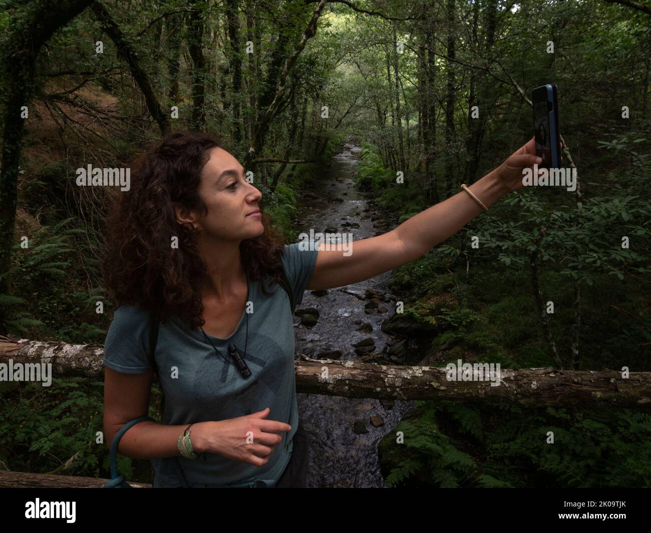 Brunette femme prenant un selfie sur un pont en bois avec rivière qui coule à travers un chêne et des châtaigniers en arrière-plan. Banque D'Images