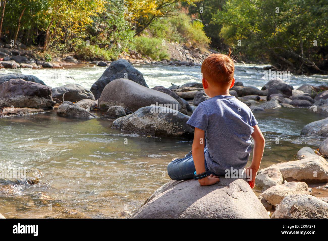 un garçon aux cheveux rouges est assis sur un rocher près d'une rivière de montagne. vue arrière Banque D'Images