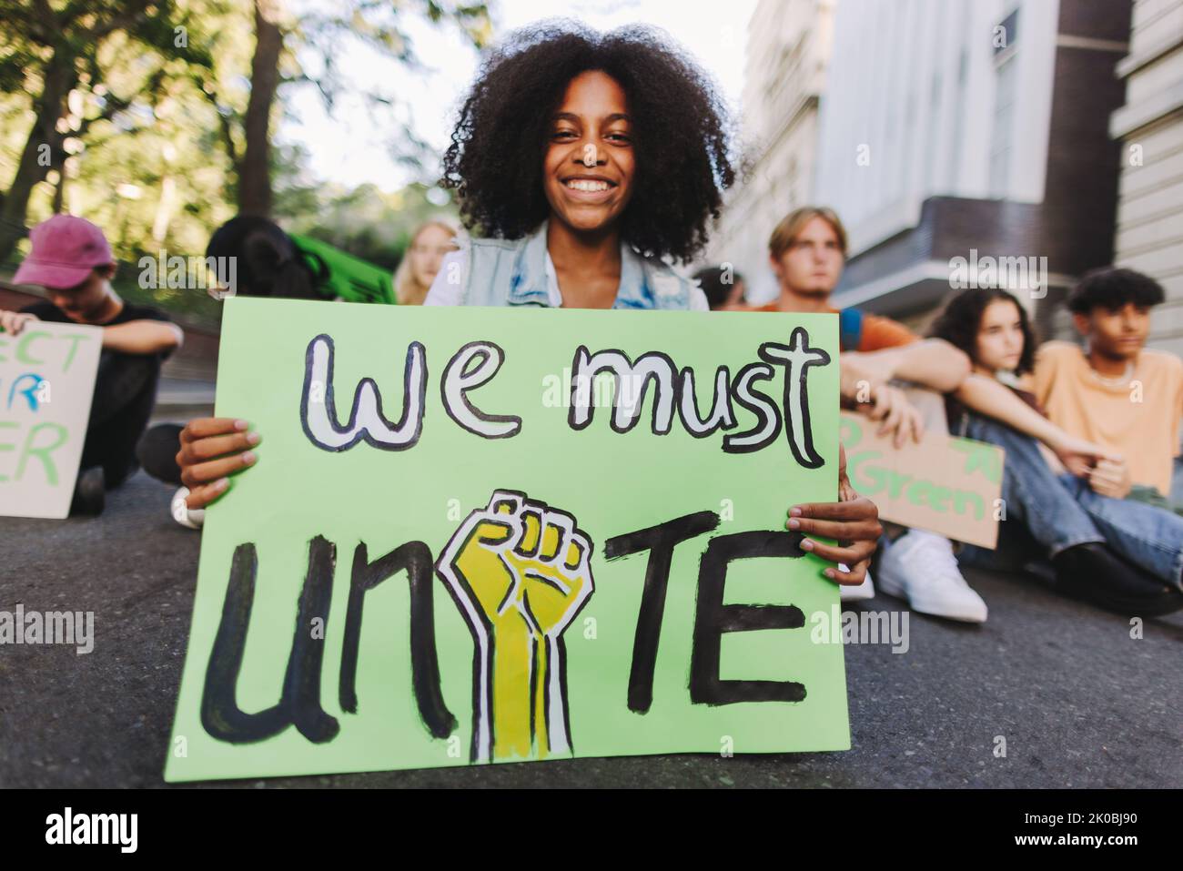 Une jeune fille noire joyeuse tenant une affiche tout en étant assise avec un groupe de manifestants lors d'une manifestation contre le changement climatique. Des jeunes militants multiculturels se joignent à TH Banque D'Images