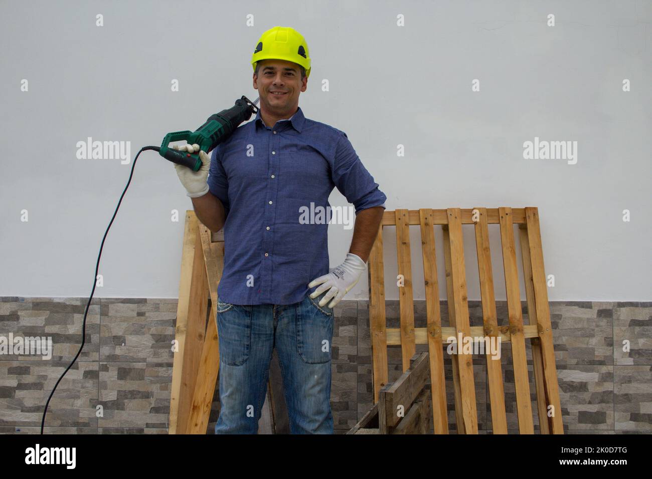 Image d'un homme de main souriant avec un casque de sécurité jaune et une scie à métaux électrique sur son épaule. Travail de coupe de bois et de plate-forme de palettes à faire soi-même Banque D'Images