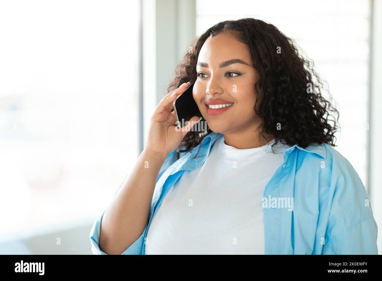 Femme noire parlant sur un téléphone portable debout près de la fenêtre à la maison Banque D'Images