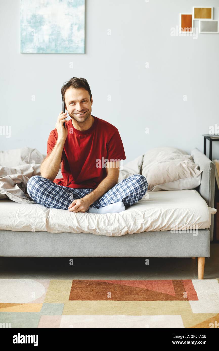 Jeune homme souriant assis sur son lit et parlant sur son téléphone portable le matin dans sa chambre Banque D'Images
