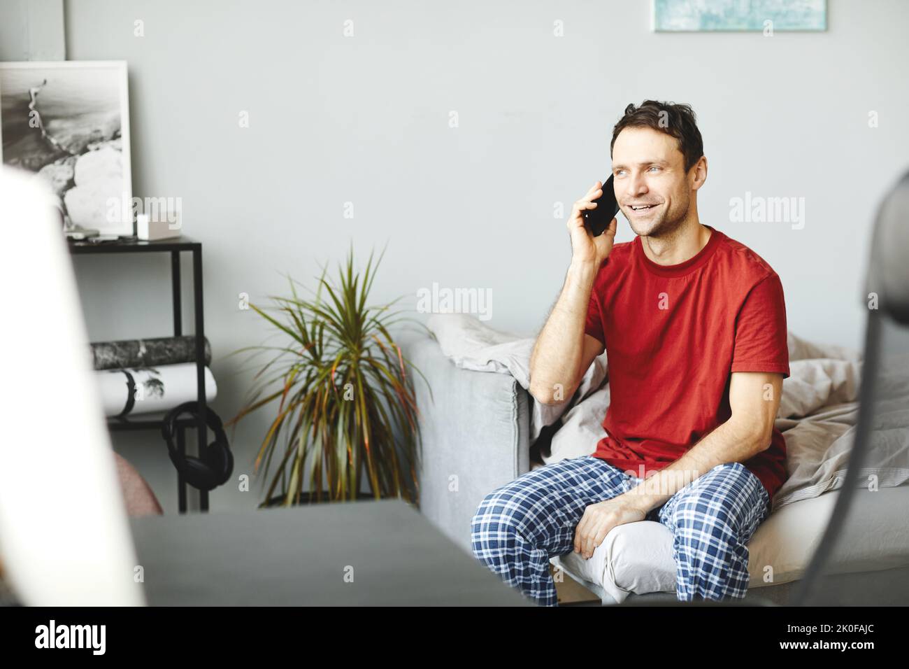 Jeune homme qui a la conversation sur téléphone portable tout en étant assis sur le lit dans sa chambre Banque D'Images