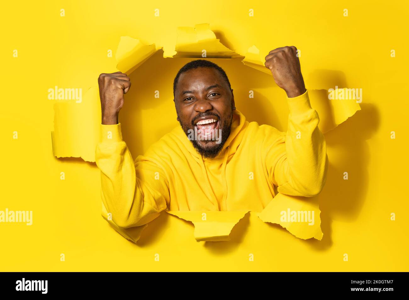 Un homme afro-américain enthousiaste qui rafle les poings, célébrant le succès et le sourire, posant dans un trou jaune déchiré Banque D'Images