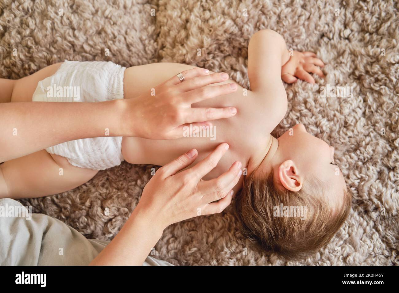 Femme mère effectuant un massage du dos à un bébé heureux allongé sur un couvre-lit beige. Maman masseuse et jeune garçon souriant. Enfant âgé d'un an et deux mois Banque D'Images