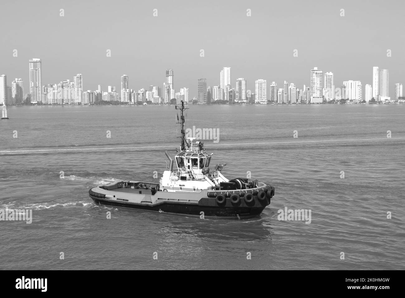 Un bateau à remorqueurs sur une mer calme contre des bâtiments de haute hauteur, en échelle de gris Banque D'Images