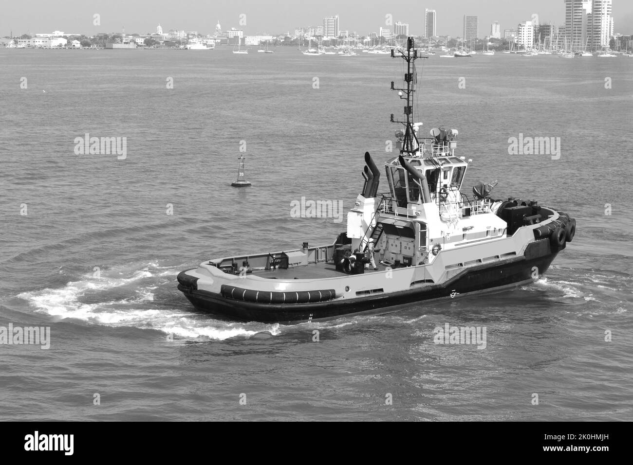 Un bateau à remorqueurs sur une mer calme contre des bâtiments de haute hauteur, en échelle de gris Banque D'Images