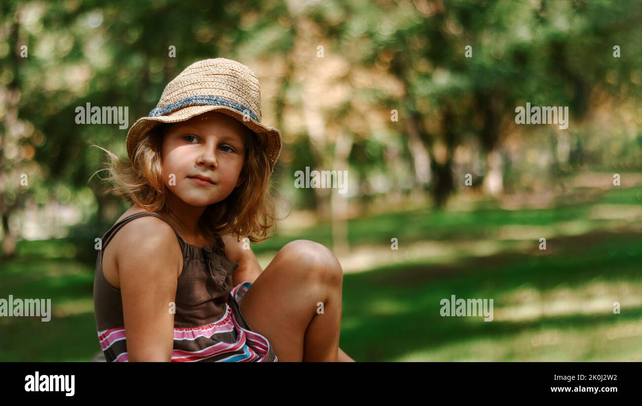 Fille dans un chapeau de paille dehors. Portrait d'une jolie fille de 5 ans. L'enfant est assis dans le parc. Copier l'espace. Banque D'Images