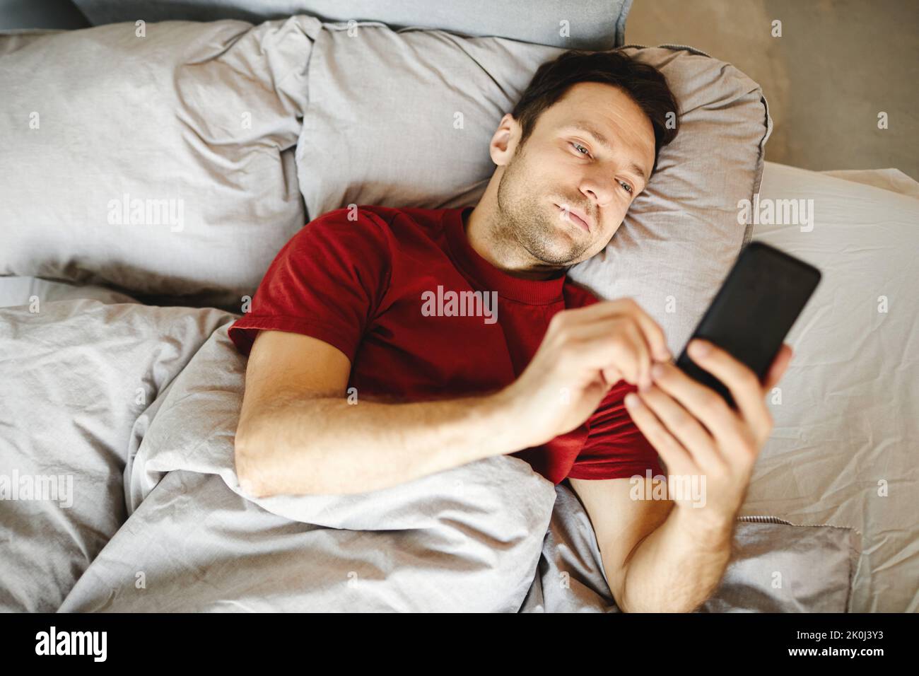 Vue en grand angle de l'homme mûr allongé sur le lit le matin et vérifiant le courrier dans son téléphone mobile Banque D'Images